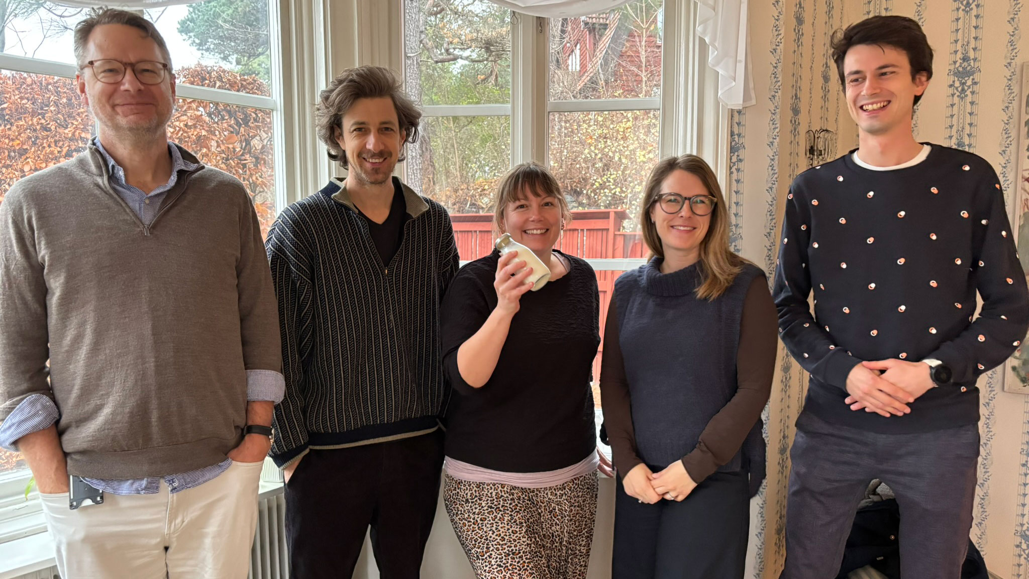 Two women and three men stand in front of a window indoors. The woman in the middle holds up a small glass bottle of milk. Photo.
