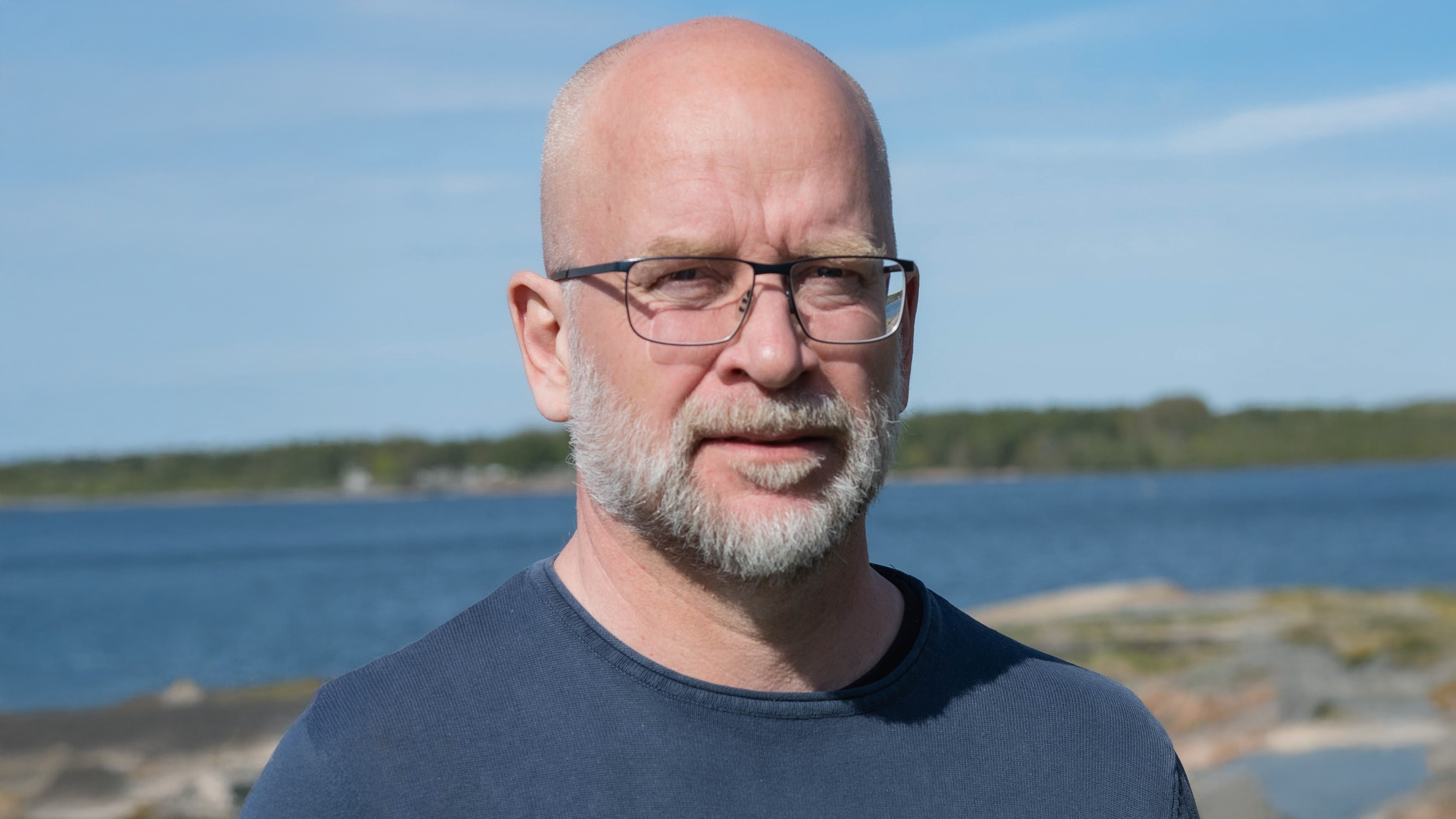 Portrait of a man wearing glasses and a beard, outdoors by the sea on a sunny day.
