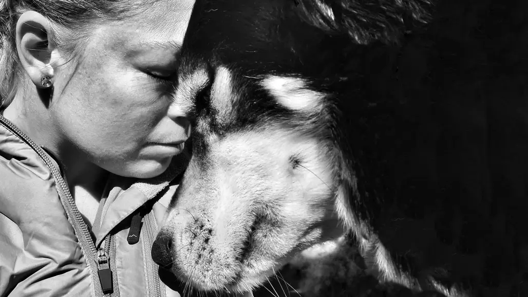 En kvinna och en hund blundar med huvuden tätt intill varandra. Svartvitt foto.