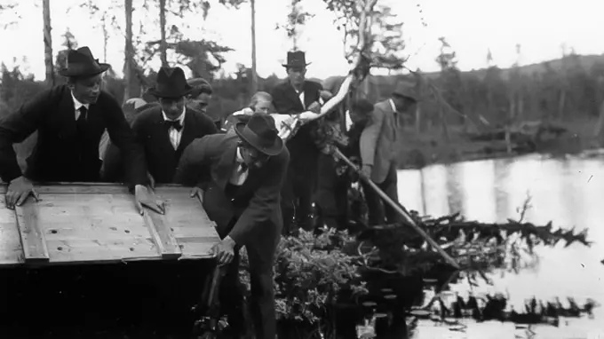 Svartvit historisk bild där flera män i kostym och hatt står vid en skogstjärn och tippar en stor trälåda mot vattnet.