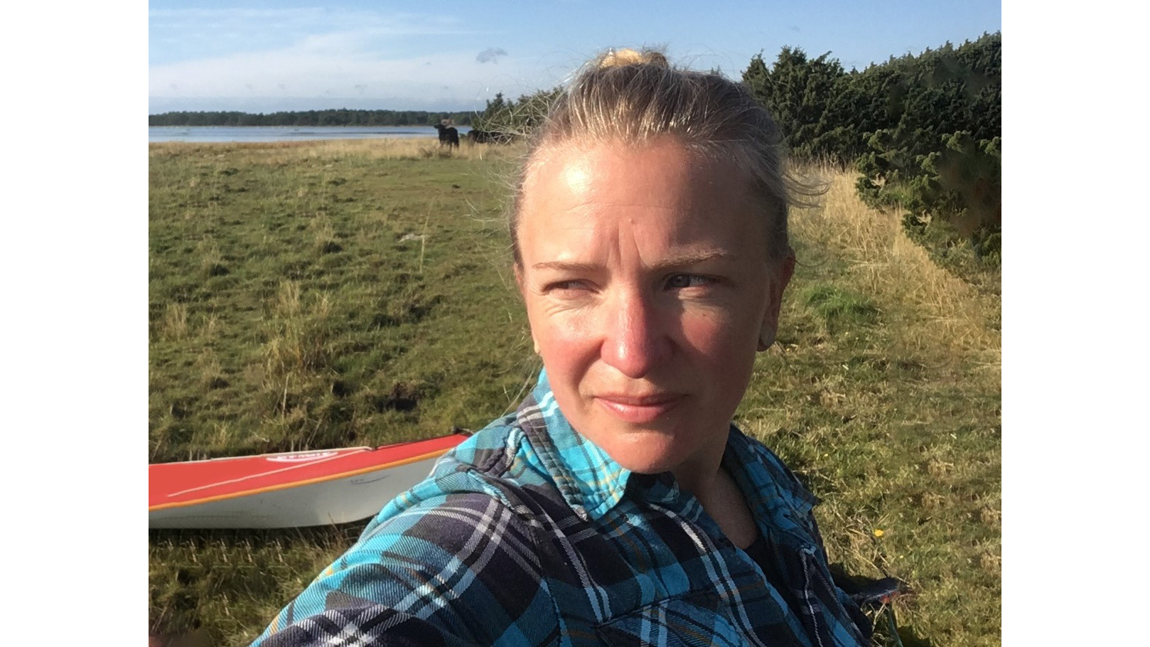 Blond kvinna med uppsatt hår ochrutig skjorta står på gräsyta vid hav eller vattendrag. Foto