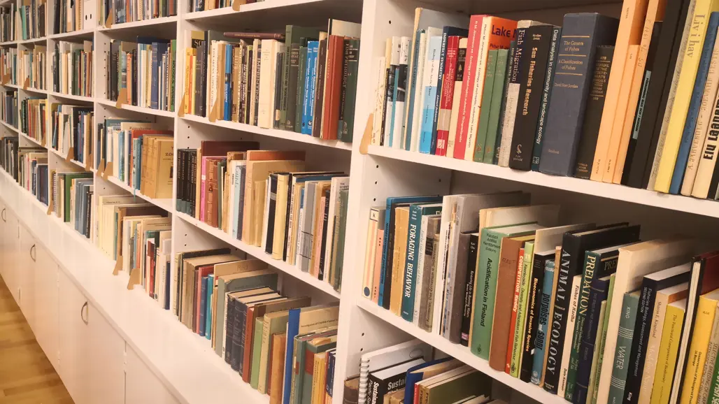 A long white bookshelf filled with non-fiction and scientific books, mainly on ecology, water management and fisheries. The books are tightly packed in different colors and sizes. The floor is light wood, and the shelf has cabinets with silver handles at the bottom.