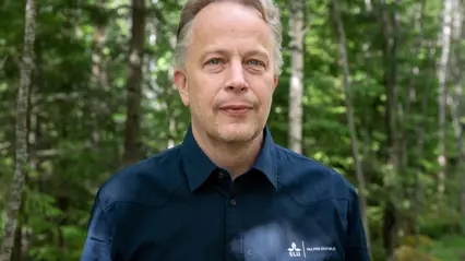 En person med kort, grått hår står i en skog med gröna träd i bakgrunden. Personen bär en mörkblå skjorta med en liten logotyp och ser mot kameran.