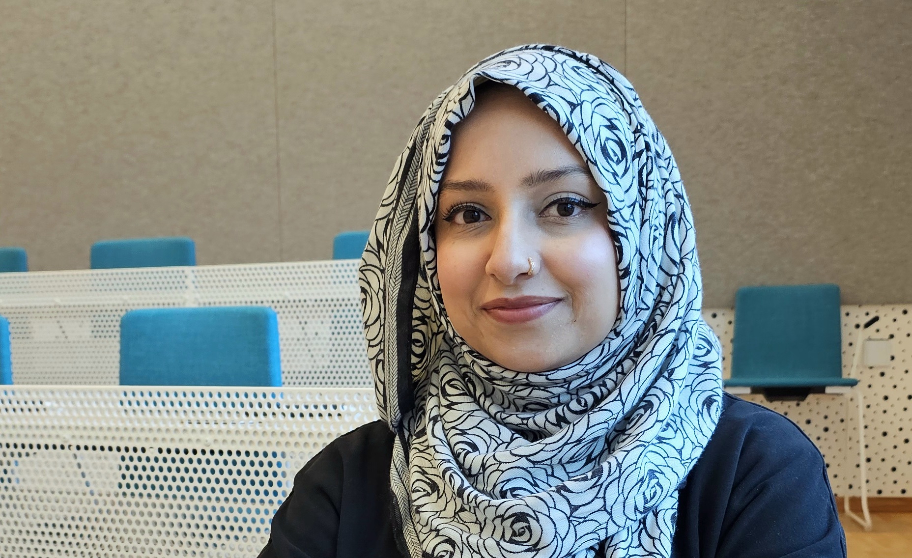 A student sitting at a table in a classroom at SLU. She is smiling towards the camera. In the background are chairs with blue seats and light grey walls.