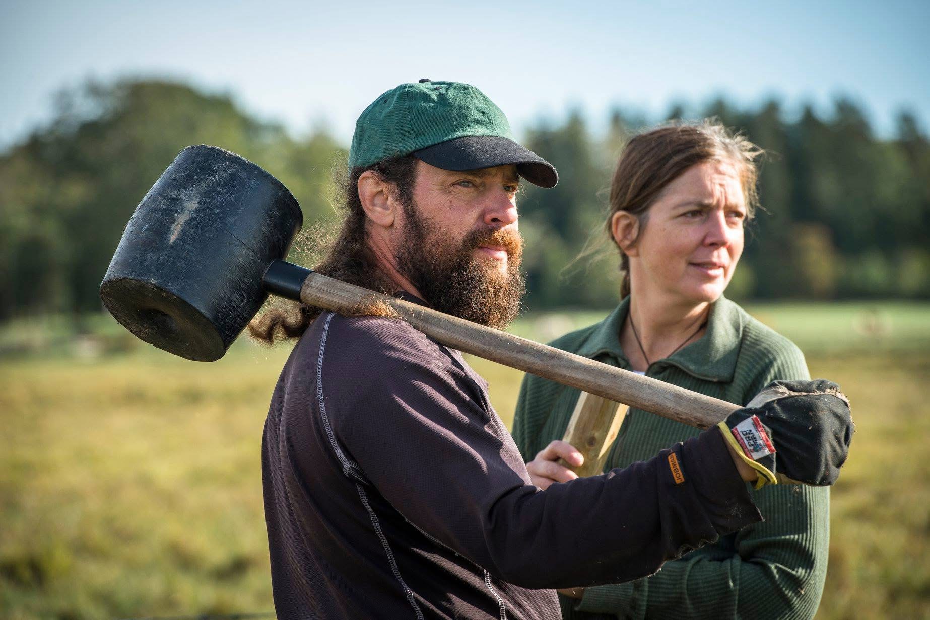 Two people are standing in a meadow, one has a large mallet over his shoulder. Photo.