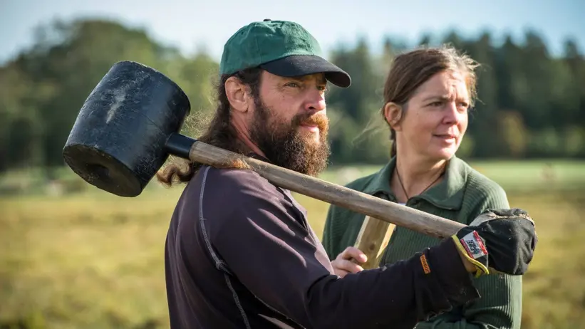 Two people are standing in a meadow, one has a large mallet over his shoulder. Photo.