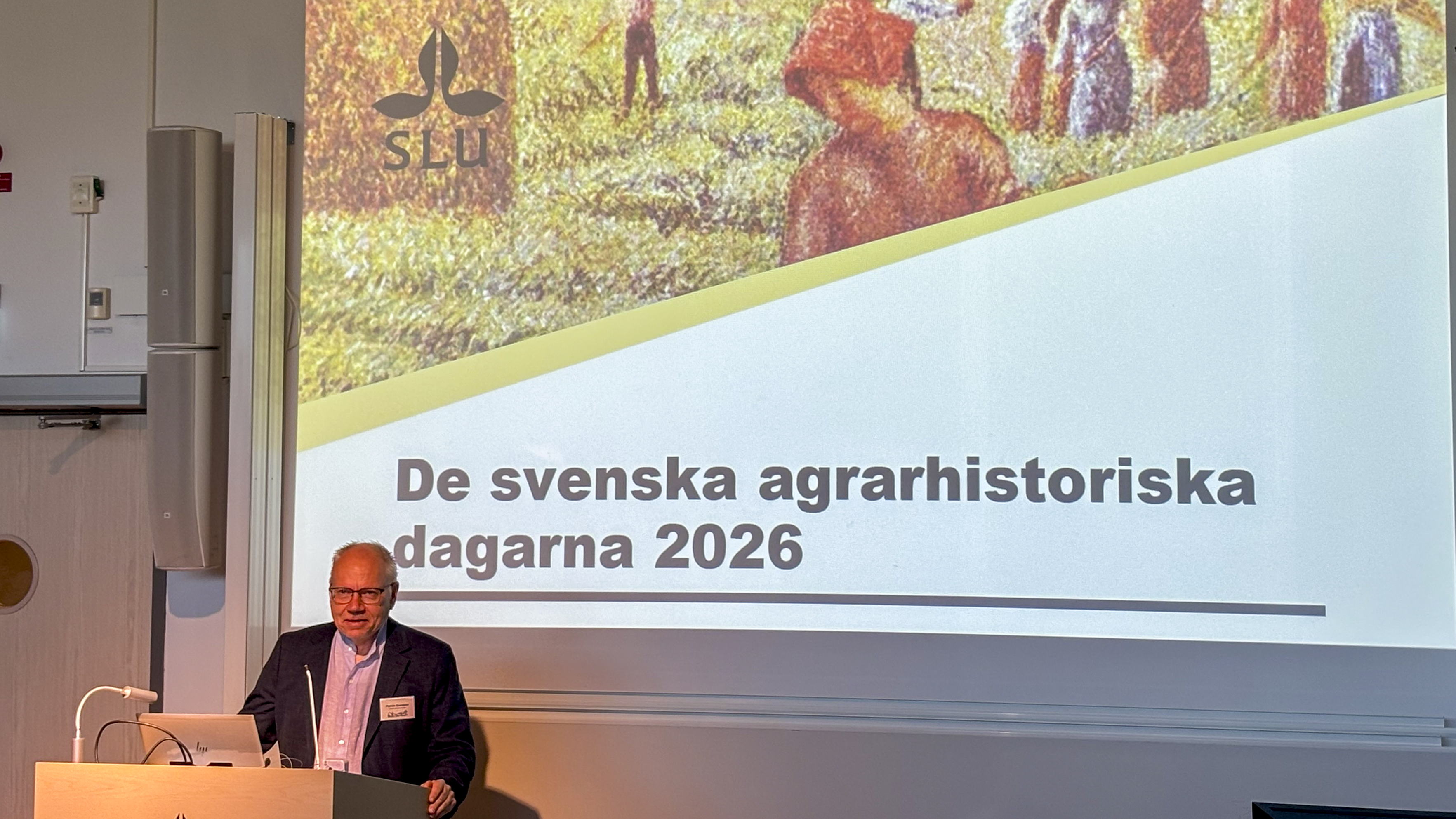 En man står vid ett podium i en föreläsningssal, en presentation i bakgrunden visar texten "De svenska agrarhistoriska dagarna".