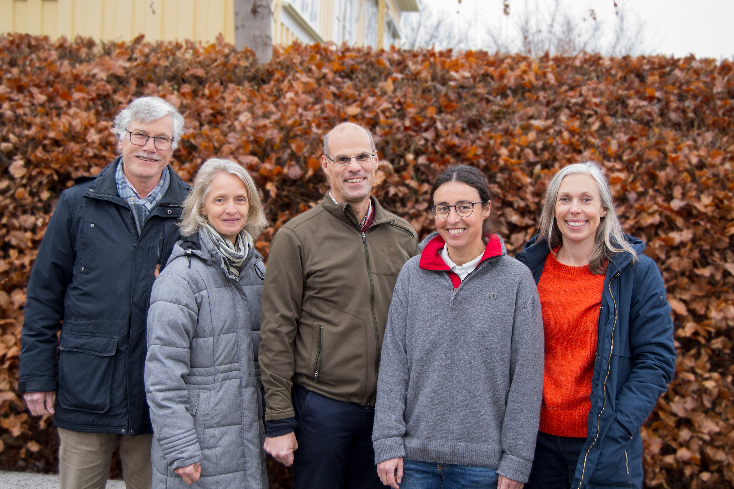 Five persons in front of a shrubbery in autumn colors. Photo.