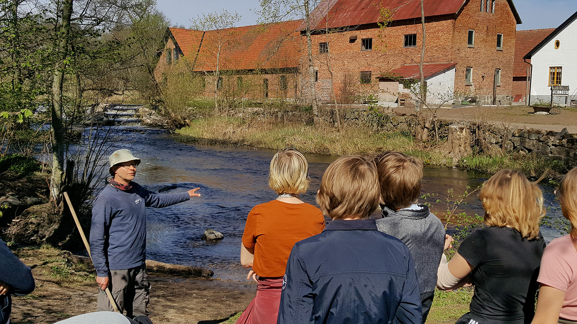 En man står framför en grupp elever vid ett vattendrag. Han håller en håv. I bakgrunden syns en gammal tegelbyggnad.