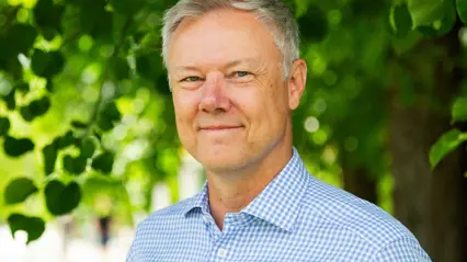 En person med kort, grått hår står utomhus med gröna löv i bakgrunden. Personen bär en ljusblå, rutig skjorta och ser vänligt mot kameran.