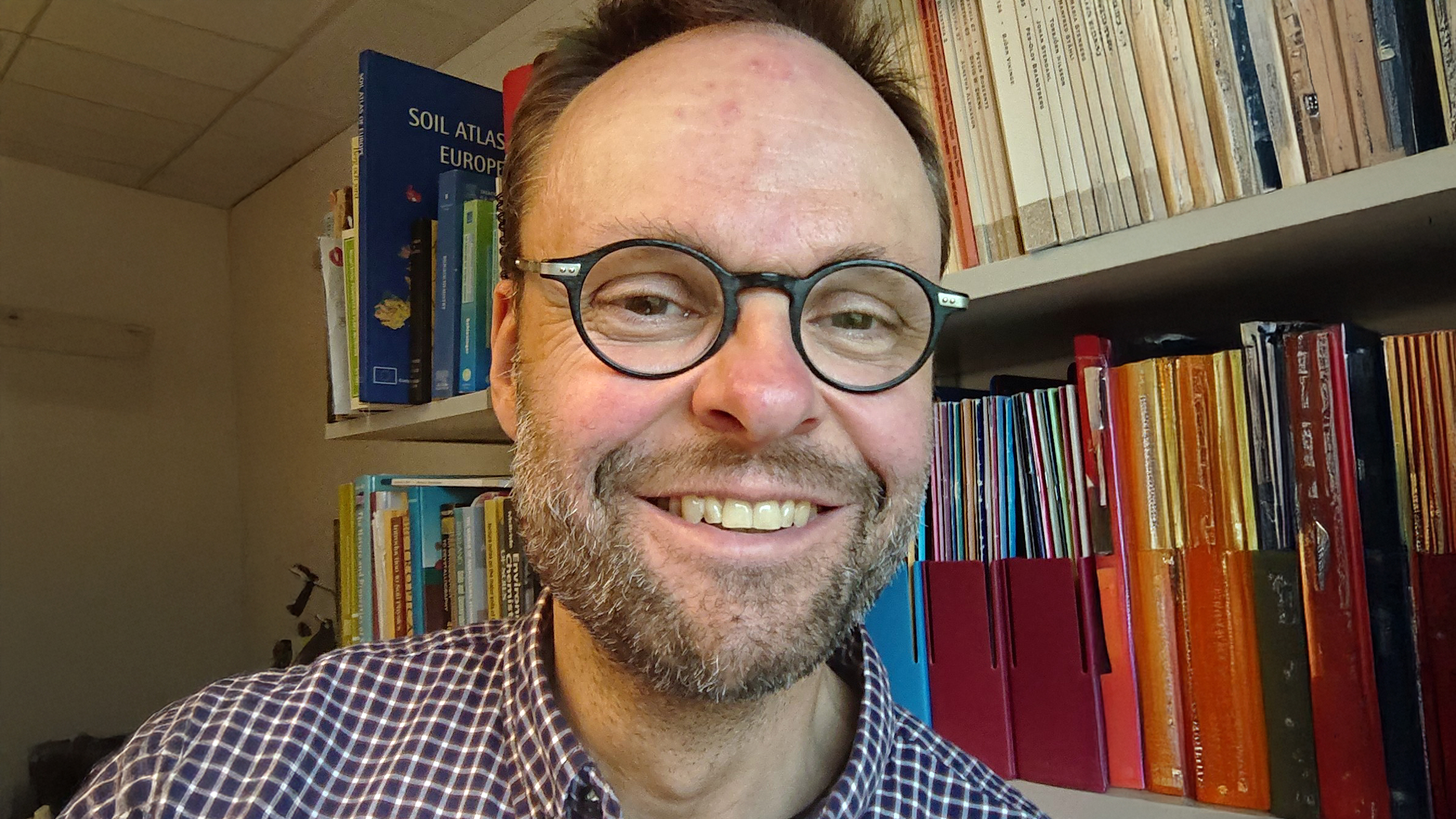 Portrait of a smiling person with glasses indoors in front of a bookshelf.