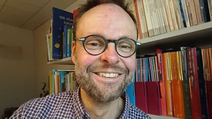 Porträtt av leende person med glasögon inomhus framför bokhylla.