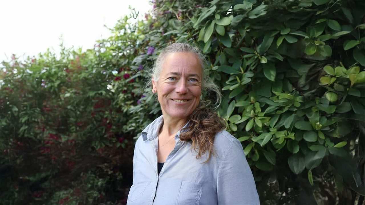 A smiling woman with long hair tied back in a ponytail stands outdoors in front of a leafy bush.