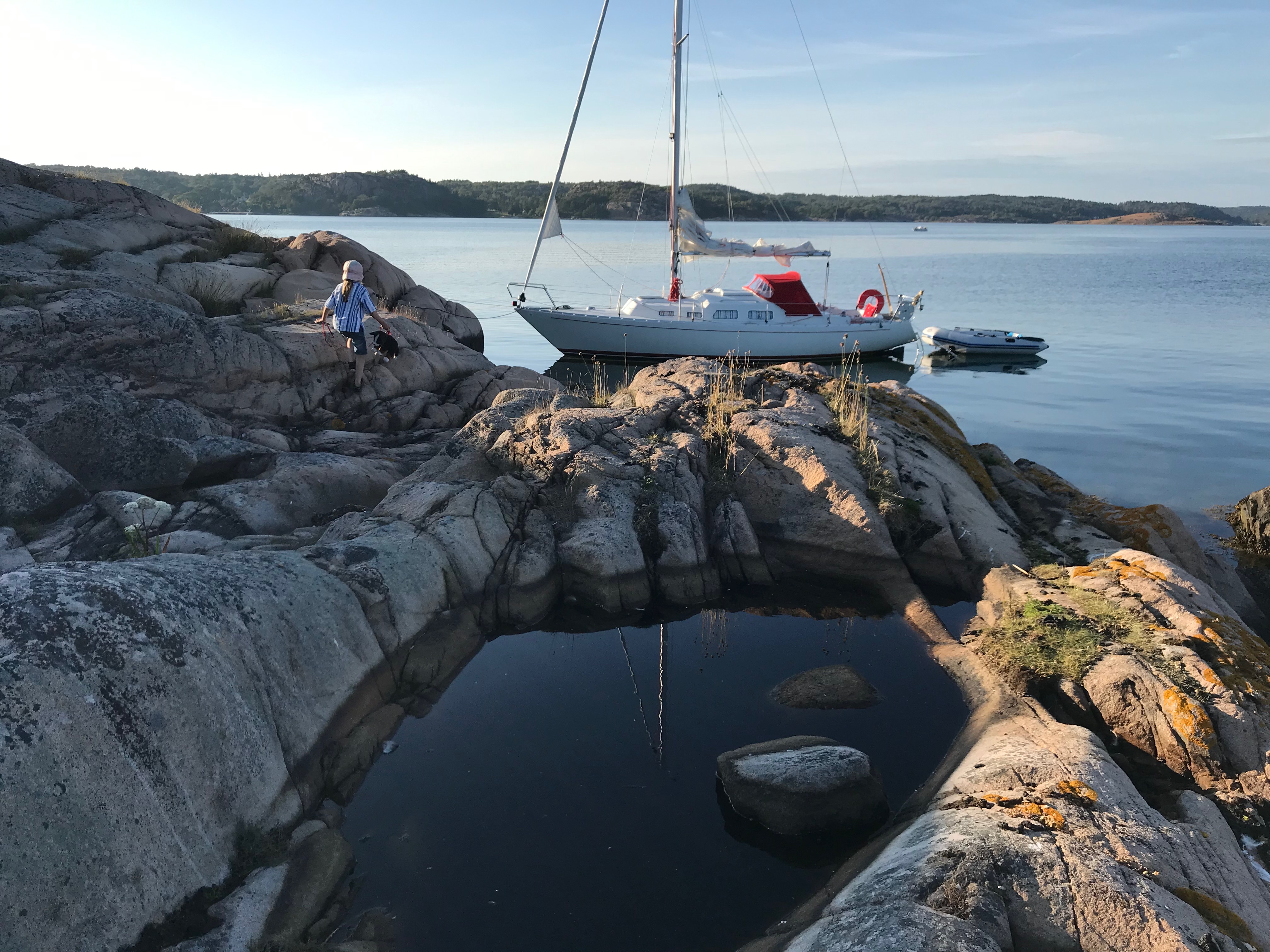 En segelbåt ligger för ankar vid en solig klippa i skärgården. Ett barn går över klipporna mot båten. 
