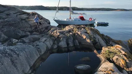 En segelbåt ligger för ankar vid en solig klippa i skärgården. Ett barn går över klipporna mot båten.