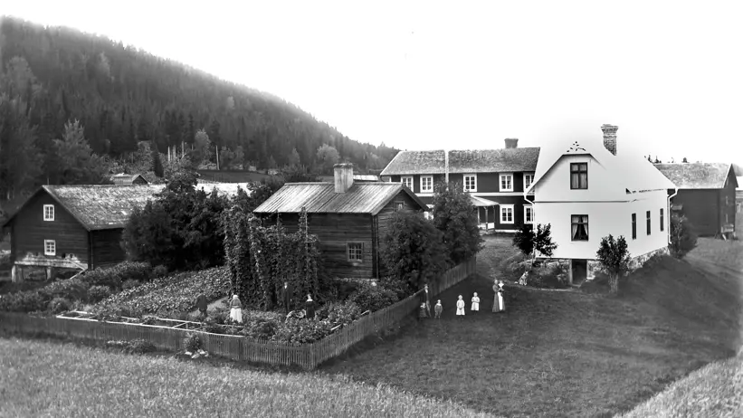Ett svartvitt foto visar en gård i Jämtland. Gården består av bostadshus och lador. Framför huset står familjen och tittar på fotografen.