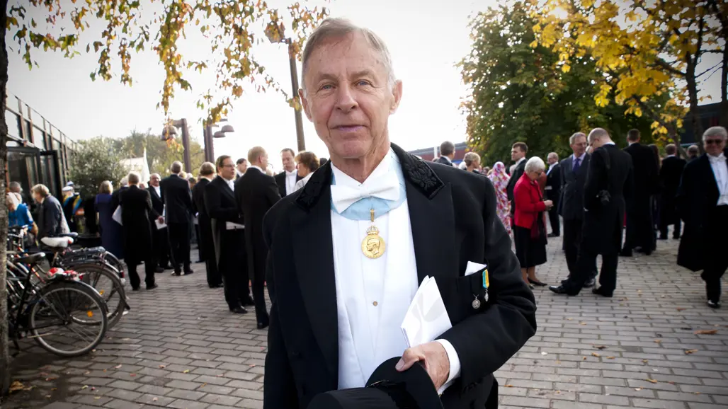 En äldre man iklädd frack och med en guldmedalj runt halsen håller i en doktorshatt.