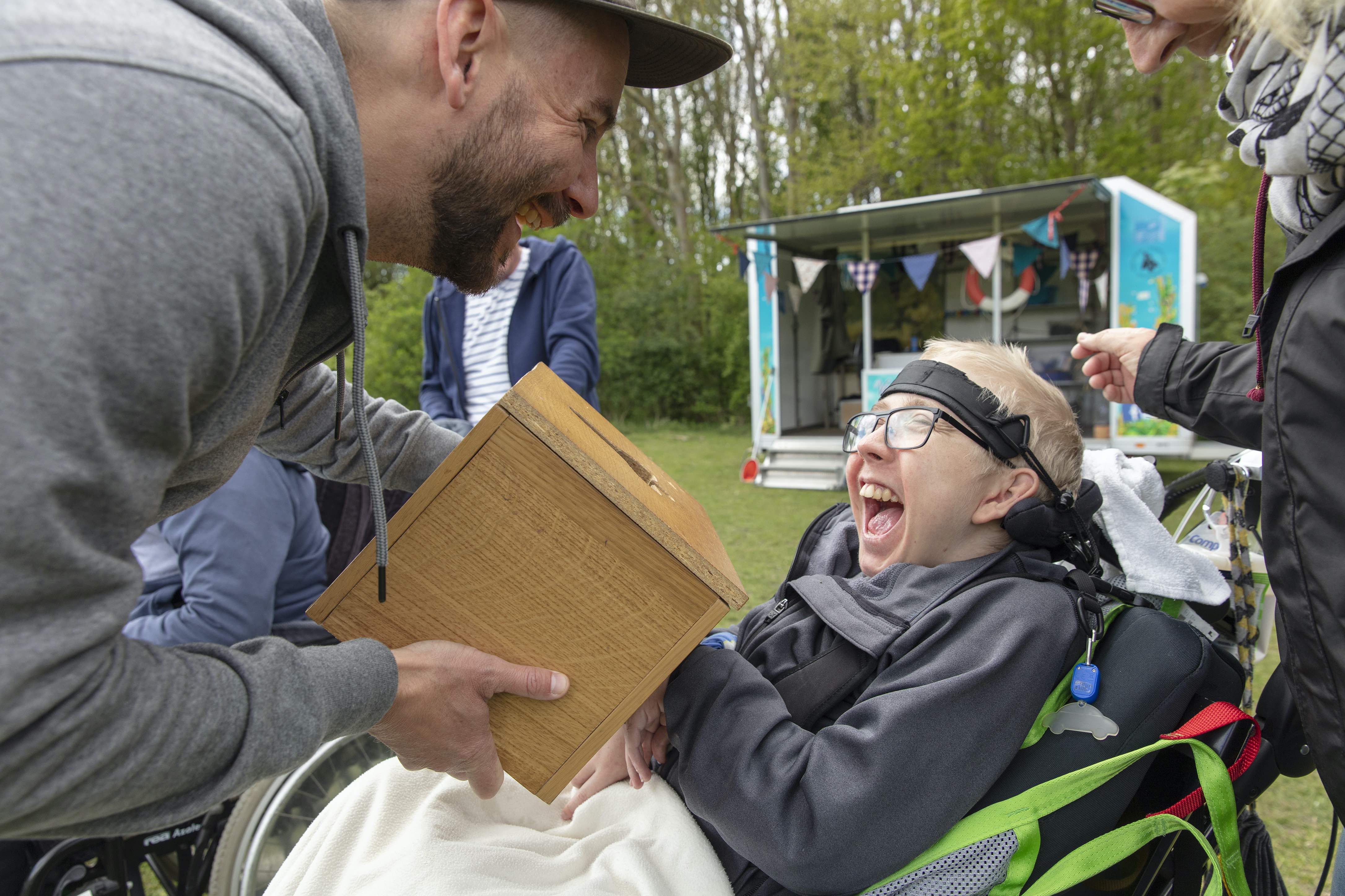 Ett skrattande barn sitter i rullstol och får lukta på något i en burk. 