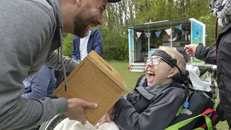 Ett skrattande barn sitter i rullstol och får lukta på något i en burk.