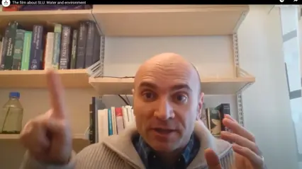 En man sitter i ett kontor framför en bokhylla och pratar engagerat mot kameran. Han pekar uppåt med ena handen och håller upp den andra som i en förklarande gest. I bokhyllan bakom honom syns facklitteratur och en flaska med vätska.