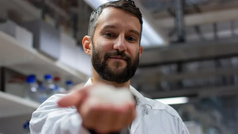 Photo of a man in a lab coat (Benjamin Schmuck, SLU) reaching out his hand toward the camera to show a piece of artificially spun spider silk.