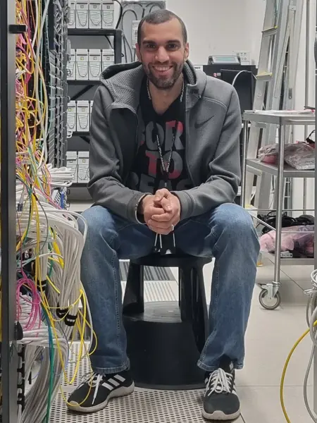 A man sits on a chair in a server hall with a lot of wires and looks happily into the camera