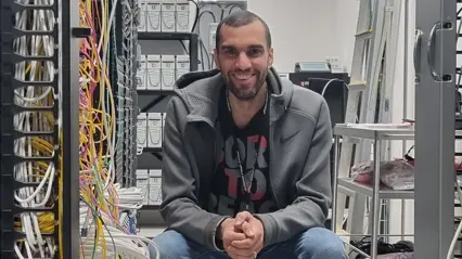 A man sits on a chair in a server hall with a lot of wires and looks happily into the camera