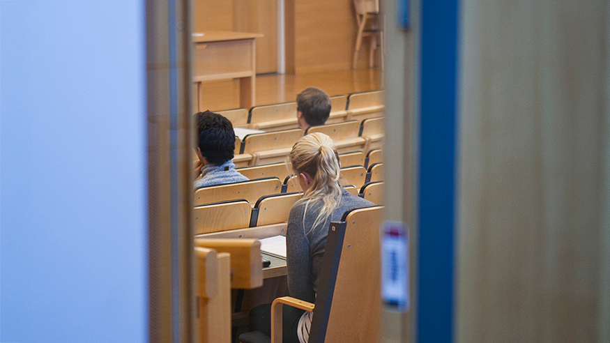Genom halvöppen dörr skymtar studenter i universitetsaula