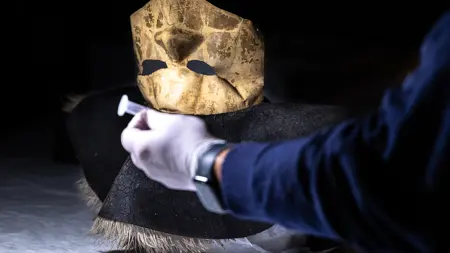 En person med handskar vid en sprucken, uråldrig guldmask som sitter fast i en mörk filthatt.