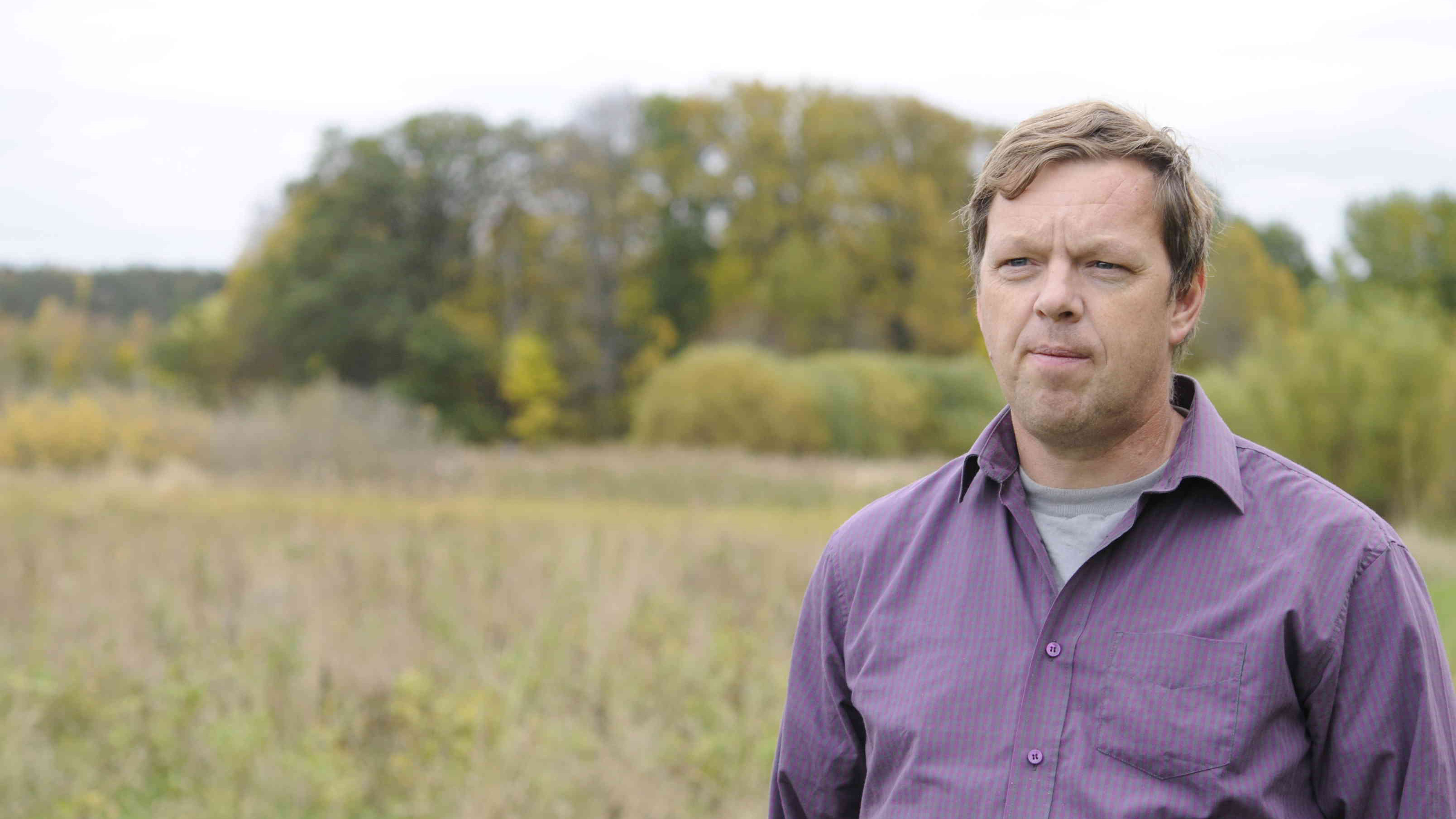 Korthårig man med lila skjorta står på fält. Foto