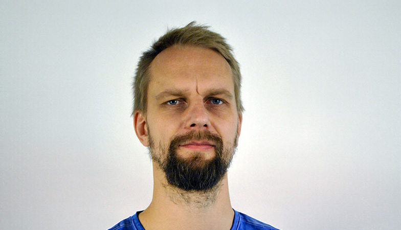 Närbild av en person som tittar rakt mot kameran. Personen har kort hår, markerat skägg och mustasch och bär en blå tröja. Bakgrunden är ljusgrå och jämnt belyst.