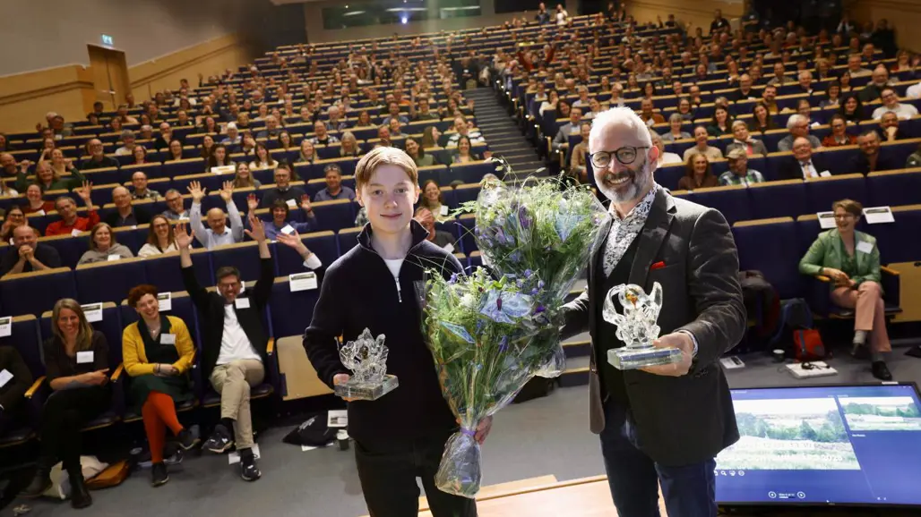 En ung man och en äldre man står i en aula med blommor och pris i händerna. Foto