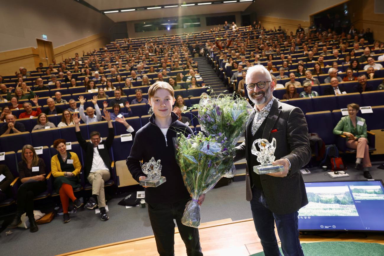 En ung man och en äldre man står i en aula med blommor och pris i händerna. Foto