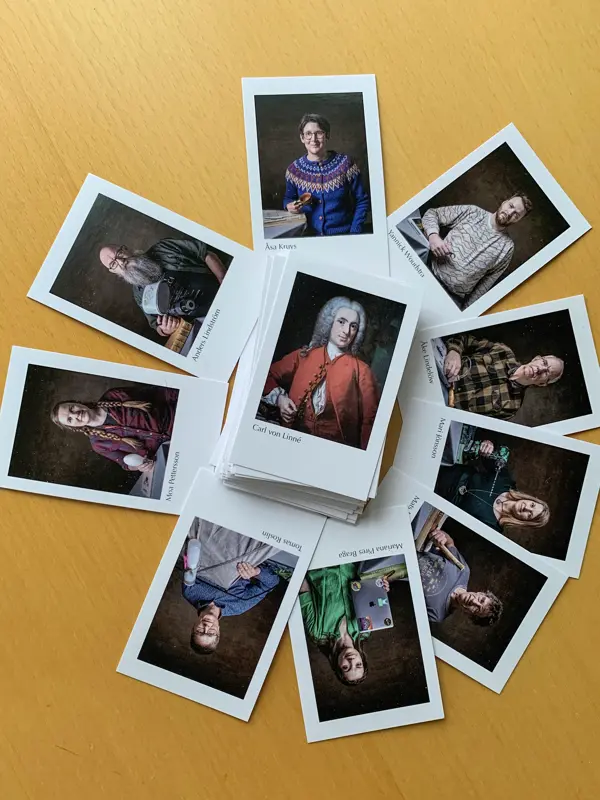 En hög med samlarbilder ligger på ett träbord, med flera bilder utspridda runtom. På korten syns porträtt av olika personer, och överst i mitten ligger ett kort med ett klassiskt porträtt av Carl von Linné.