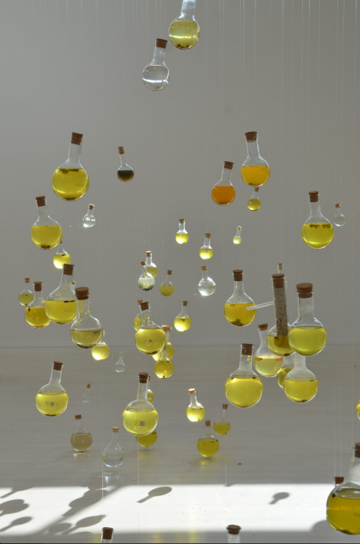 Bilden visar glasbehållare med olikfärgat vatten som hänger nerför taket. Ljuset ger skuggor av behållarna. 