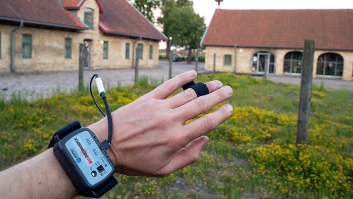 A hand with measuring instruments in front of brick buildings. Photo.