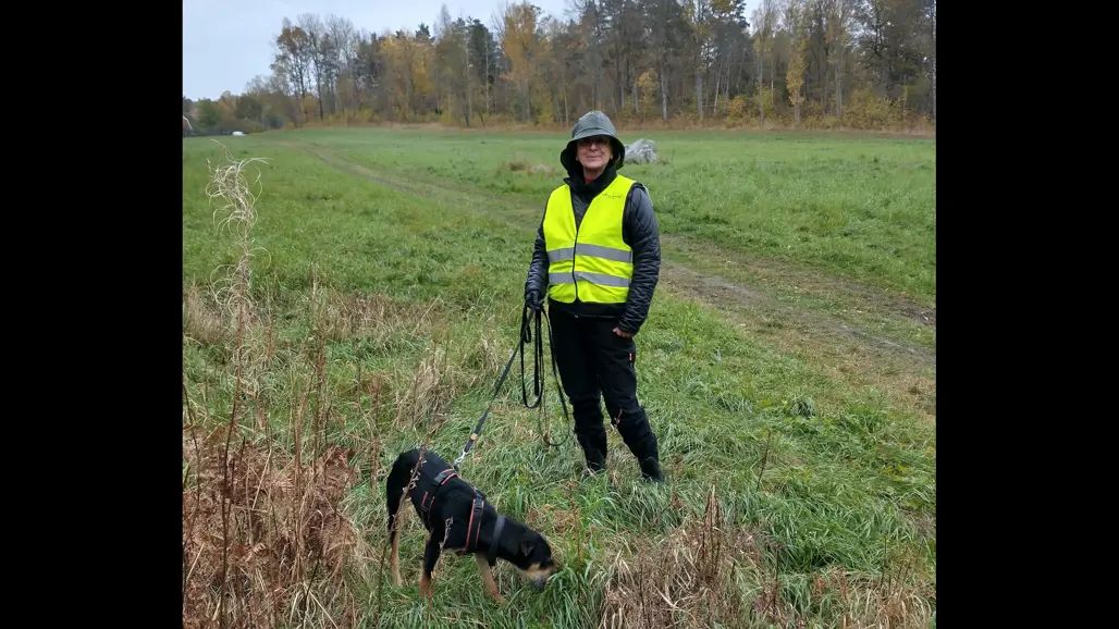 En kvinna med regnkläder och gul varselväst står på ett fält och håller en hund i koppel. Foto