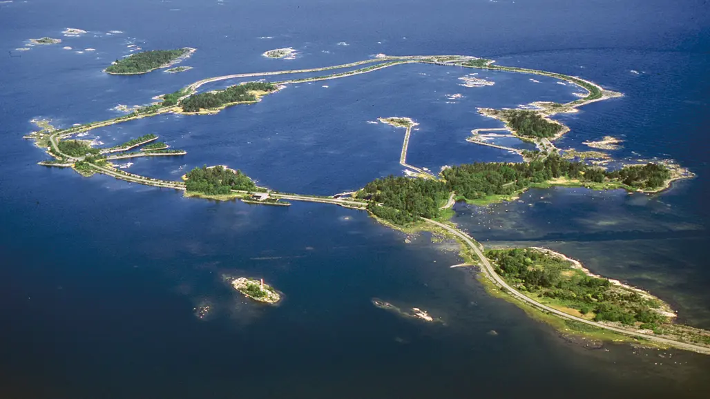 Aerial view of a coastal landscape with several interconnected islands surrounded by blue sea. Narrow roads and bridges connect the islands, which are covered in green vegetation and have small buildings and jetties. Smaller islands and islets are visible scattered in the surrounding water.