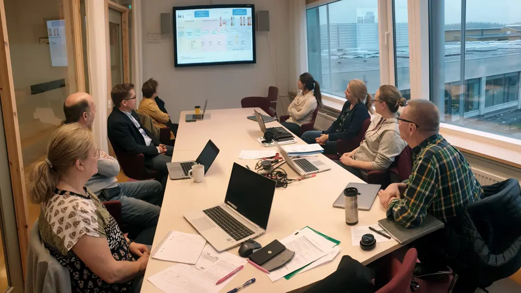 Åtta personer sitter runt ett bord och tittar på en skärm på väggen. Foto.