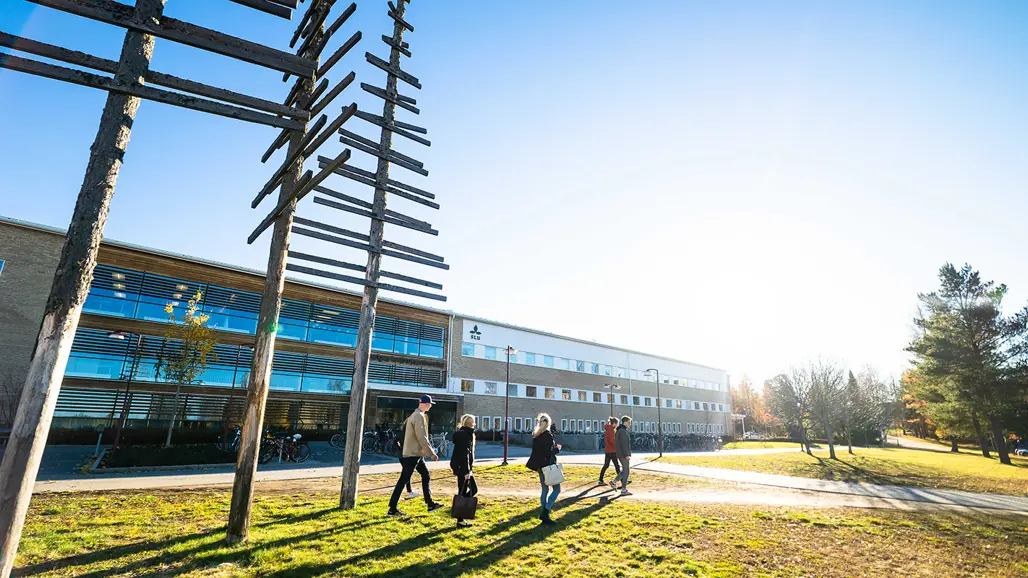 A modern building with large glass windows and several people walking on a lawn in front of it. To the left, there is an art installation of tree trunks with horizontal branches. The background features trees and a clear sky with sunshine.