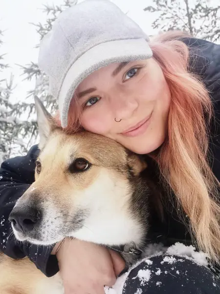 En person i vinterkläder och keps ligger i snön och kramar en hund, med snötäckta träd i bakgrunden.