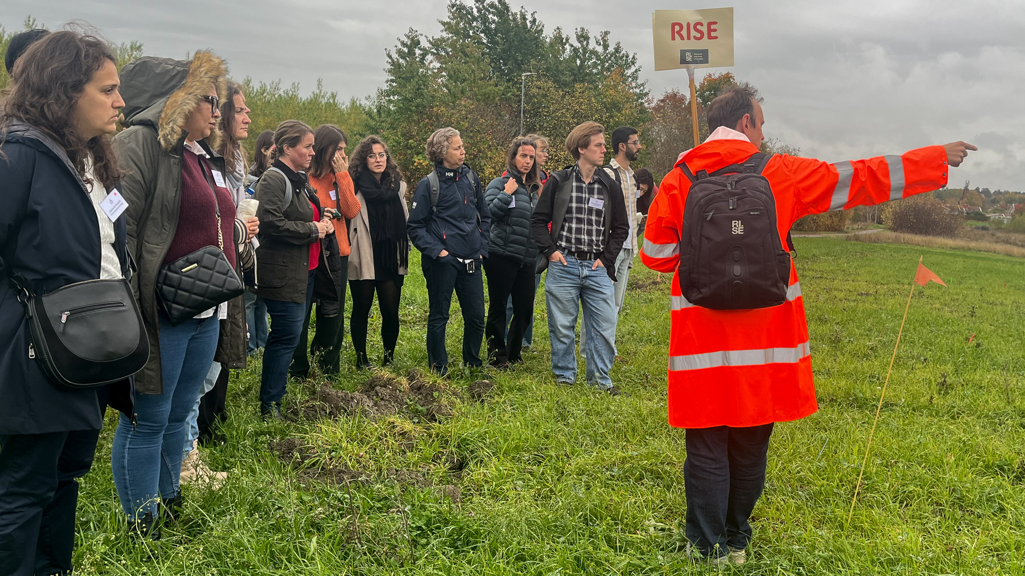 En person i orange jaca står framför en grupp människor på ett fält. Foto.