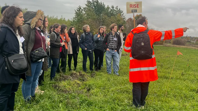 En person i orange jaca står framför en grupp människor på ett fält. Foto.