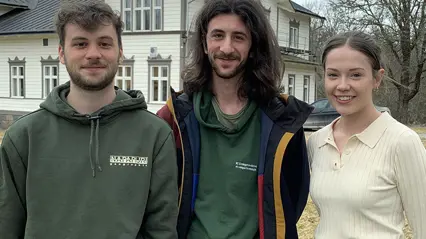 Halvkroppsbild på tre unga människor som står framför ett vitt hus, två män och en kvinna. Foto.