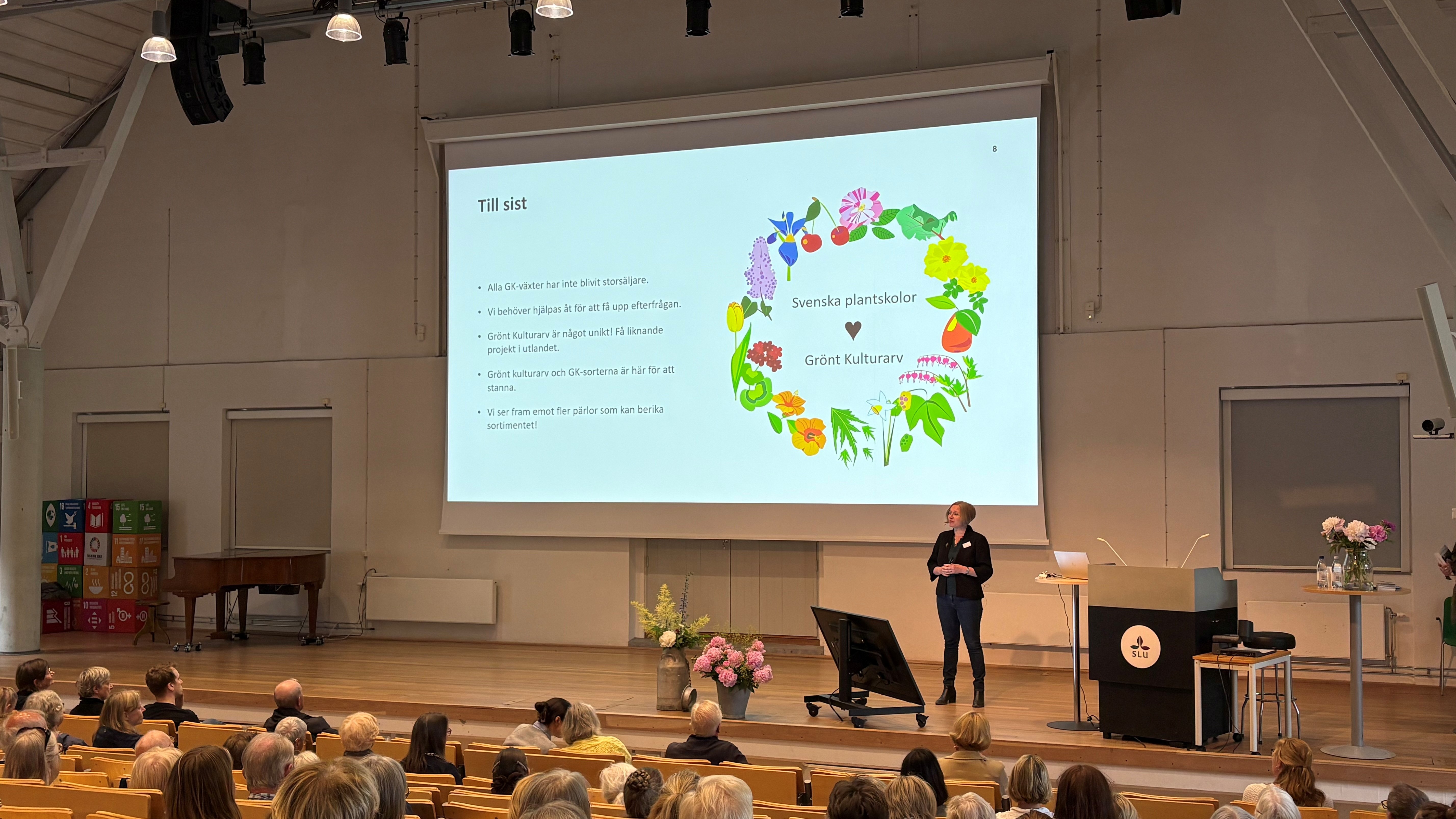 Helena Jauhiainen står på en scen och framför en stor skärm. På skärmen står "Svenska plantskolor älskar Grönt kulturarv".