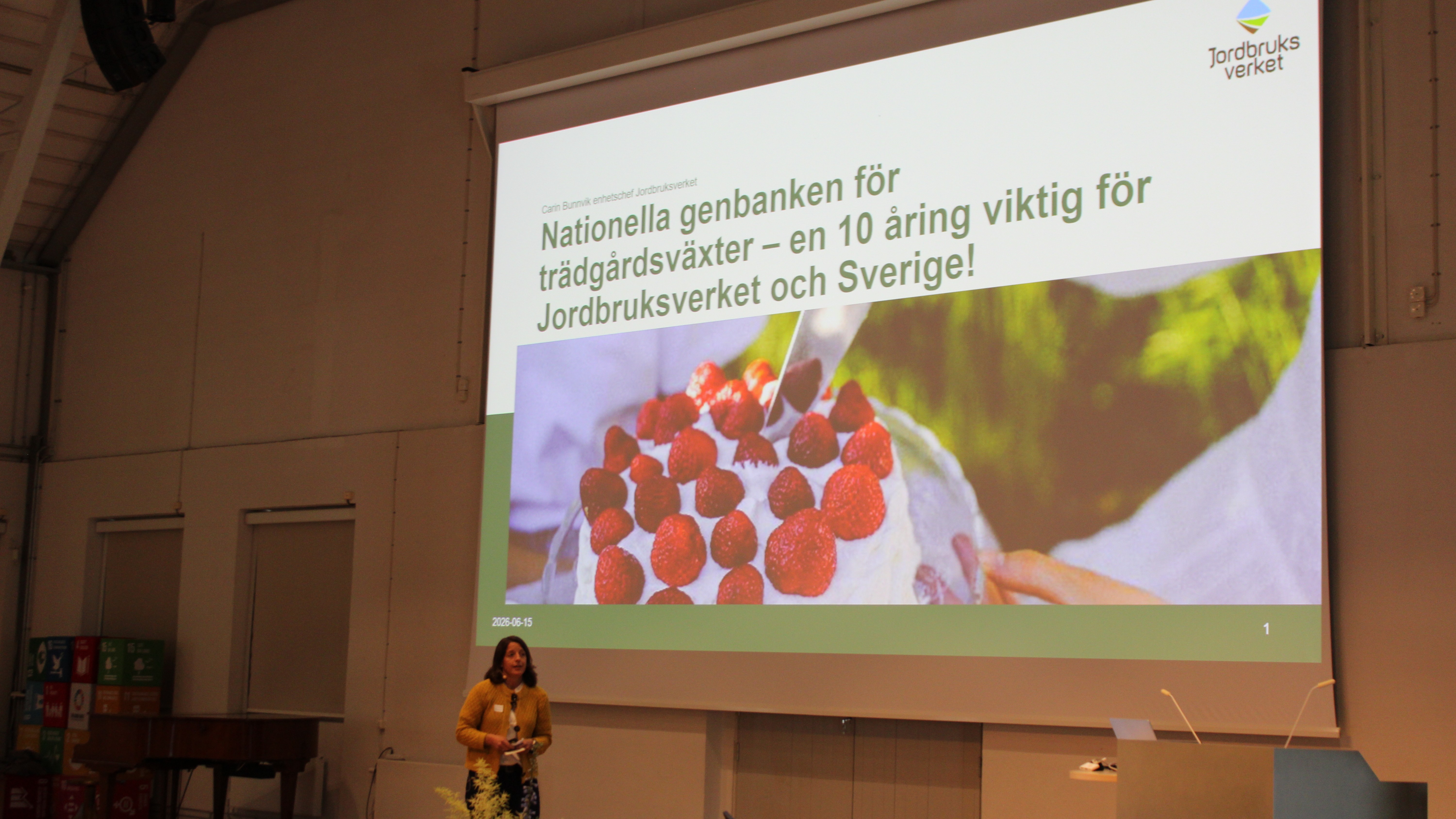 Karin Bunnvik från Jordbruksverket har en presentation framför en stor skärm. På skärmen syns en stor tårta och det står "Nationella genbanken för trädgårdsväxter. En 10-åring viktig för Jordbruksverket och Sverige."