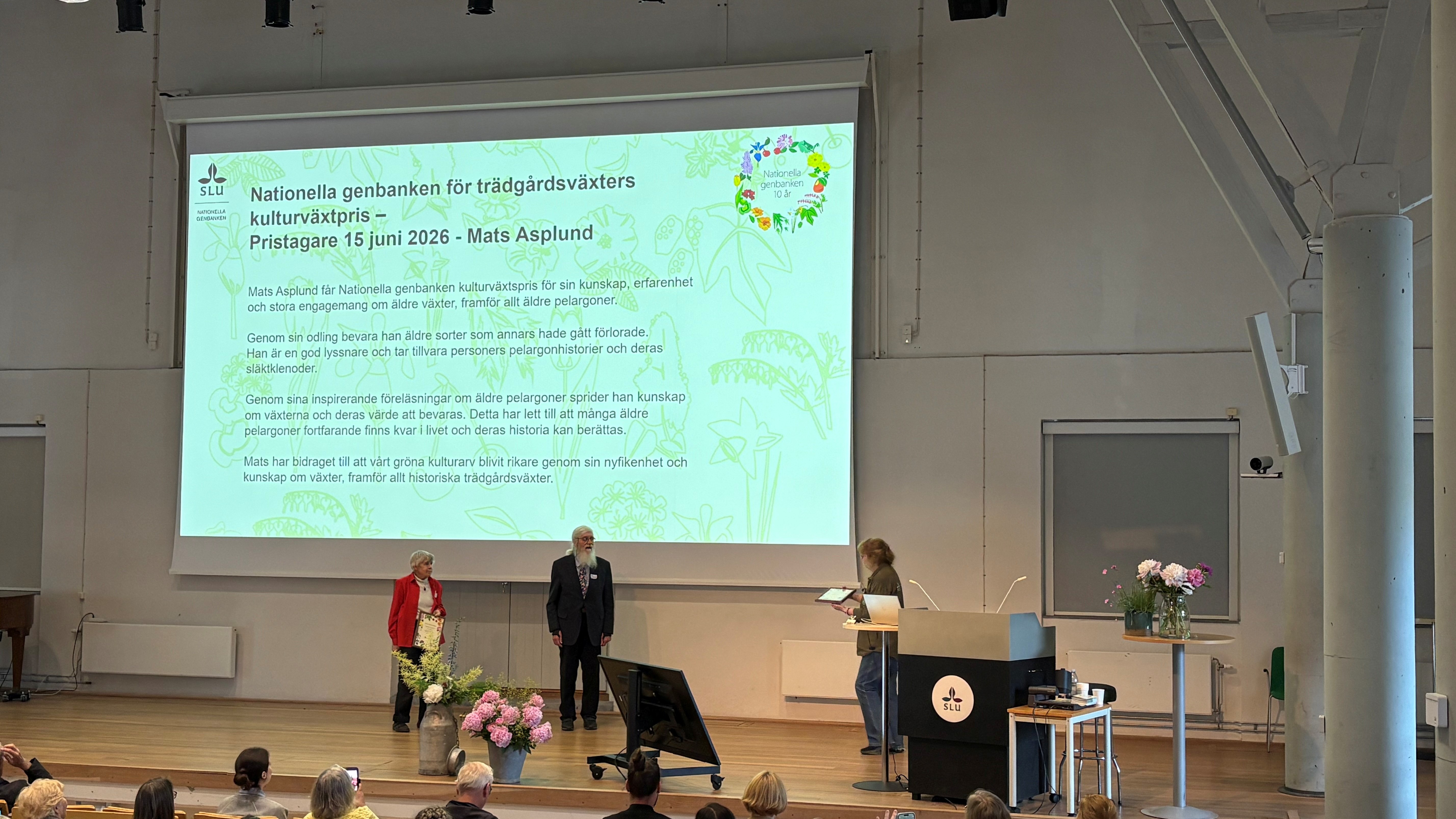 Mats Asplund står på scenen i Alnarpsgårdens aula och tar emot Kulturväxtpriset. I bakgrunden syns en stor skärm med motiveringen till priset. 