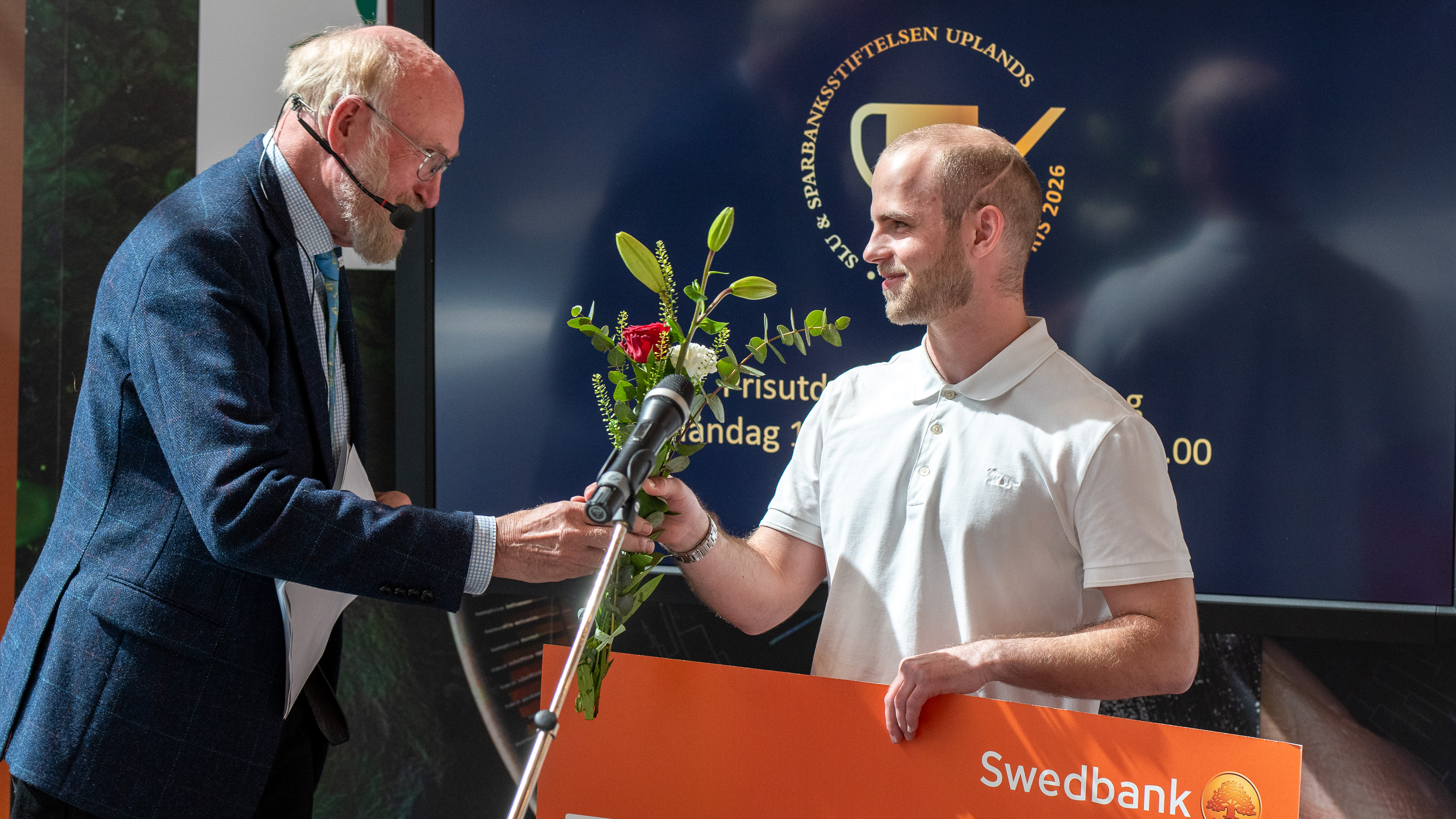 En man tar emot en blombukett från en annan man på en scen. Foto.