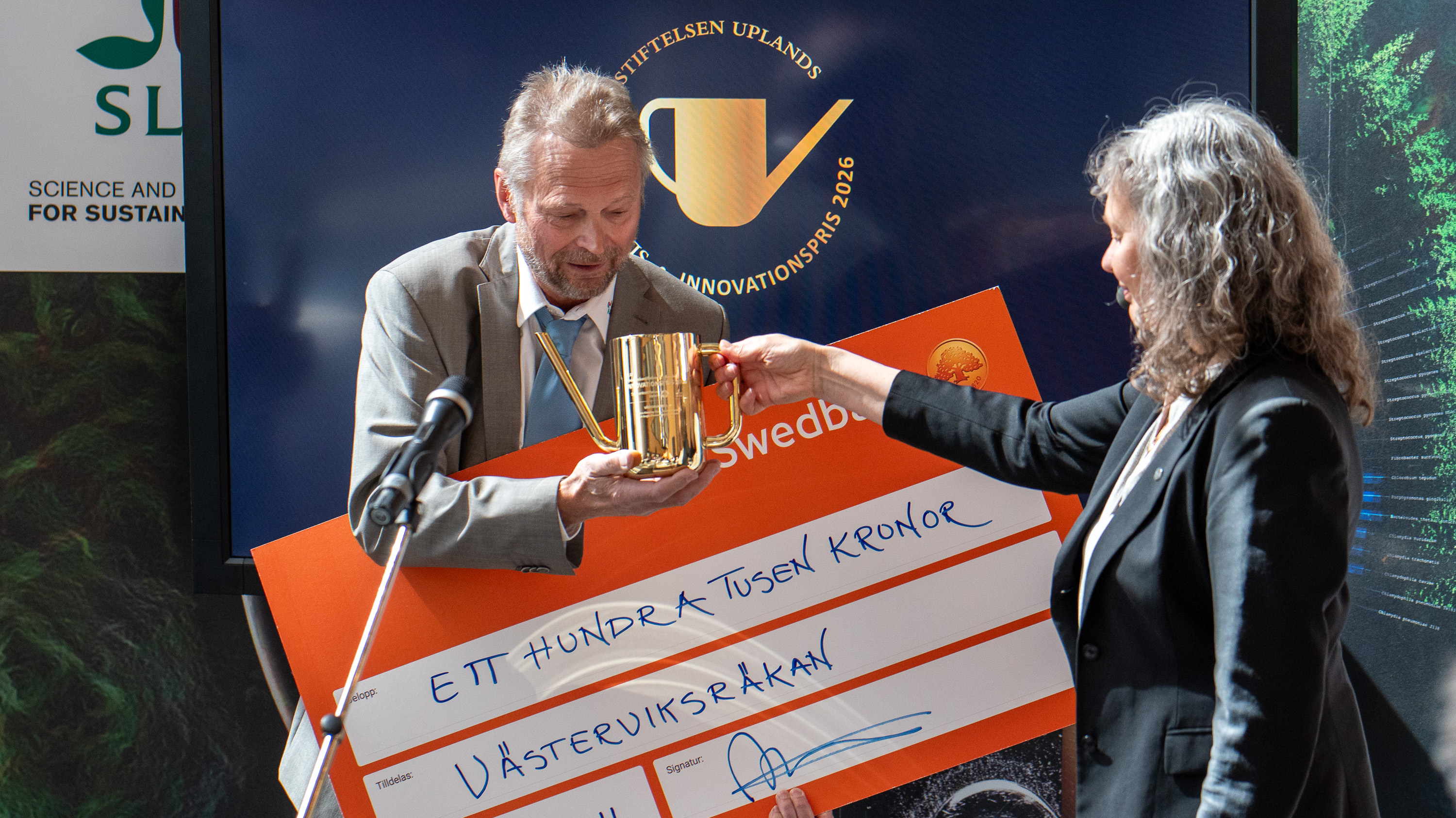 En man tar emot en vattenkanna i guld av en kvinna på en scen. Foto.