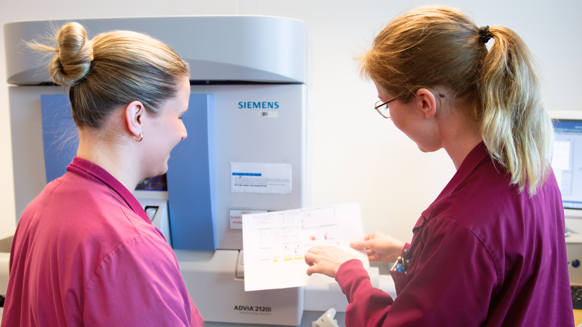 Två kvinnor med blont hår och iklädda vinröda rockar som står bredvid varandra. Kvinnan till höger sida håller i ett papper och pekar med ena handen på pappret. 